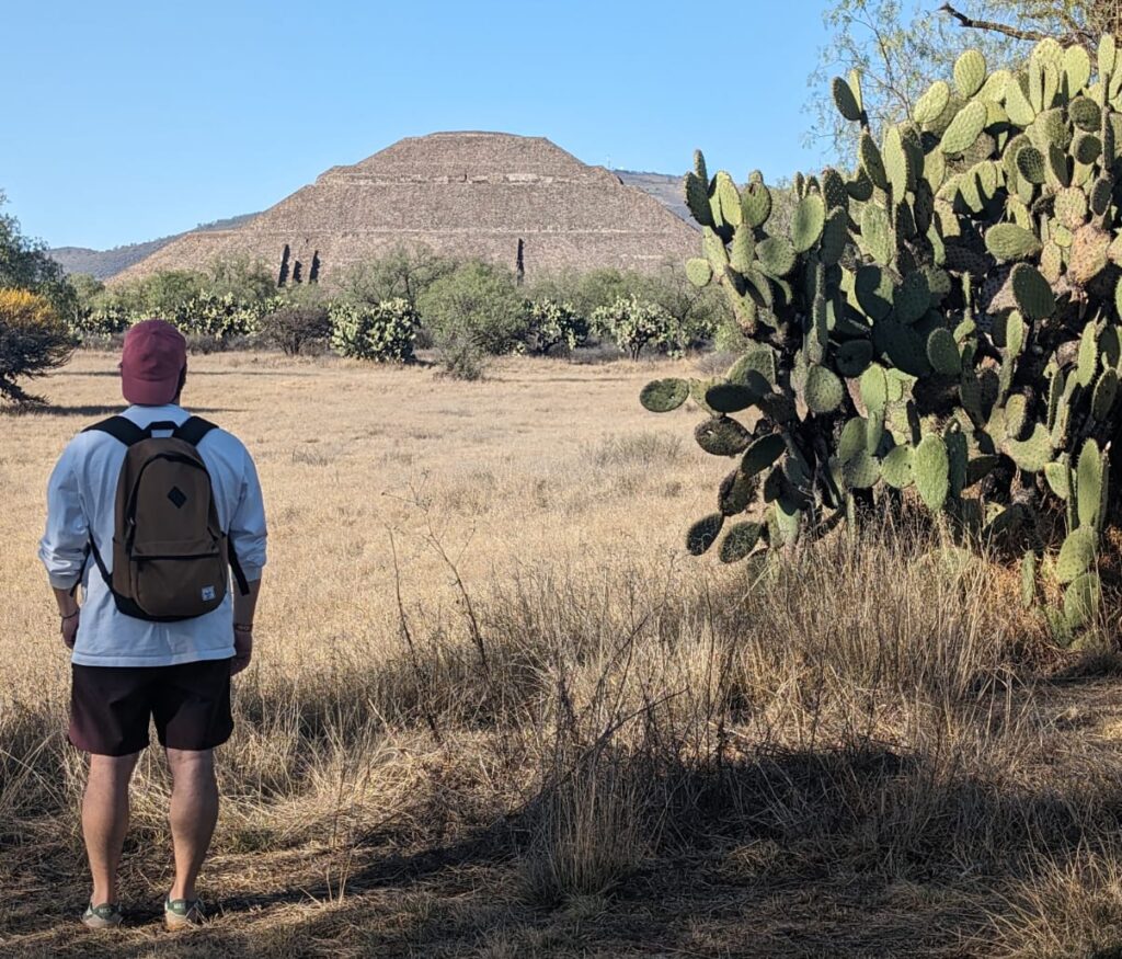 A young man stands with his back to the camera in the foreground, on the left of the frame. He is wearing dark shorts, a white sweatshirt, a red baseball cap and has a backpack on. He is looking towards some ancient ruins (pyramid) in the background. To the right of the frame is a large prickly pear plant. There is dry grass surrounding the area. There is more vegetation (bushes and small trees) in the middle of the frame.