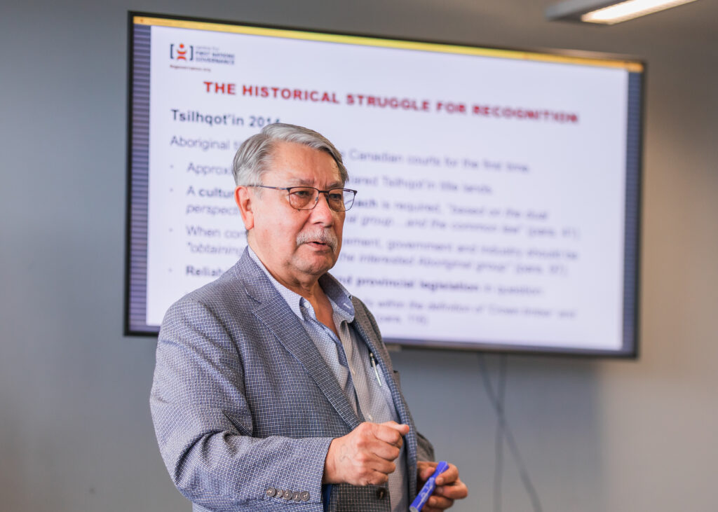 An Indigenous man wearing a grey suit jacket and a light-coloured shirt and glasses stands in front of a presentation slide that is entitled "The Historical Struggle for Recognition.” He is holding a blue marker in his left hands. His right hand is in the shape of a fist. 