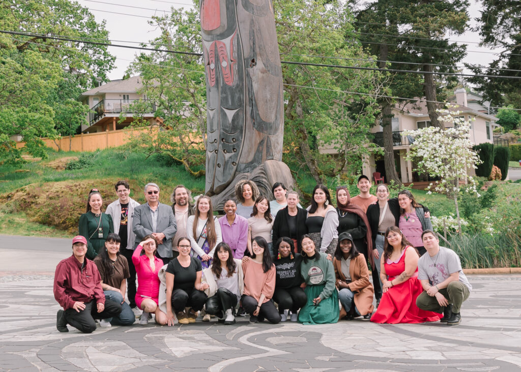 A group of 26 people of varied ages and ethnicities, some standing, some kneeling, pose for a group photo outdoors. They are standing in front of a large Indigenous wood carving. There are several trees and a couple of single family homes in the background.