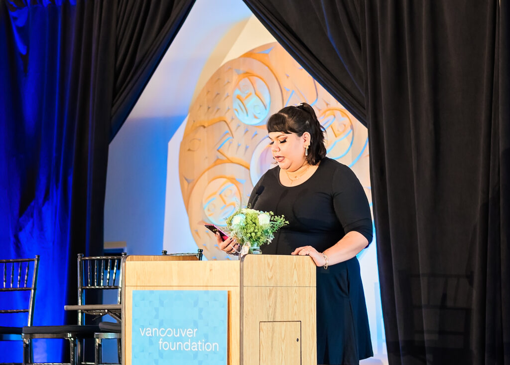 An Indigenous woman stands behind a podium with the “Vancouver Foundation” logo visible. She has shoulder-length black hair and is wearing a black dress. She’s looking down on her phone, which she is carrying in her right hand. On the podium to her right is a vase with a small bouquet of blue and white flowers. Behind her are dark blue curtains. 